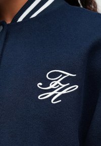 Námornícky modrá univerzitná bunda s bielymi pruhmi na golieri a bielym vyšitým iniciálmi "FH" na predstave.