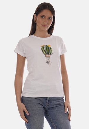 Donna che indossa una maglietta bianca con una grafica verde a forma di mongolfiera a forma di cactus con girasoli, abbinata a jeans blu, in piedi davanti a uno sfondo neutro.