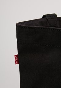 Sac en toile noire au design structuré, doté d'une étiquette rouge sur le côté et d'une surface texturée. Ouverture par le dessus sans quincaillerie visible.