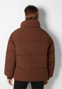 Giacca puffer marrone con colletto alto, cuciture arricciate e vestibilità comoda. Il materiale sembra morbido e leggermente opaco con un design senza cuciture.