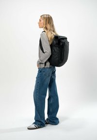 Svart ryggsäck med rulltoppdesign, justerbara axelremmar och framficka, bärs över en grå sweatshirt och lösa blåjeans.