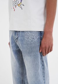 Camiseta blanca de algodón con estampado gráfico colorido, combinada con jeans de mezclilla azul claro que tienen un logo bordado en el bolsillo trasero.