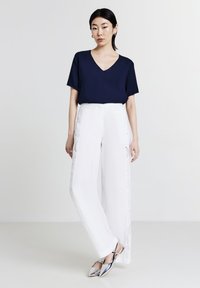 T-shirt a V scollo in blu navy abbinato a pantaloni bianchi a gamba larga con delicate applicazioni in pizzo sui lati. La modella indossa scarpe flats a punta in tonalità argentata.