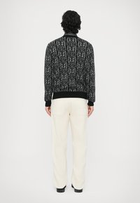 Giacca bomber nera con motivo a fantasia, dettaglio logo grigio, polsini e fascia in vita a coste, indossata con pantaloni bianchi, vista da dietro.