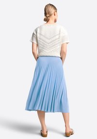 Donna con capelli biondi raccolti in un chignon indossa un top bianco a maniche corte dalla texture particolare, una gonna midi plissettata blu chiaro e sandali con tacco marroni, in piedi di spalle.