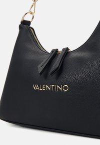Borsa a mano in pelle nera con finitura texturizzata, zip dorata e logo "VALENTINO". Presenta un manico superiore singolo e due accentuazioni a fiocco.