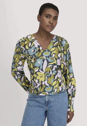 Frau trägt eine langärmlige V-Ausschnitt-Bluse mit grünem, blauem und weißem Blumendesign, kombiniert mit hellblauen Jeans, steht vor einem einfarbigen Hintergrund.