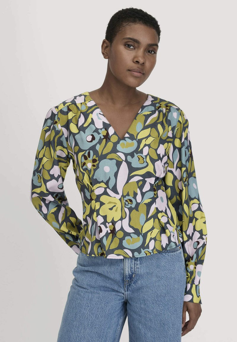 Donna che indossa una blusa a maniche lunghe con scollatura a V, decorata con un motivo floreale verde, blu e bianco, abbinata a un paio di jeans azzurri chiari, in piedi davanti a uno sfondo neutro.