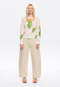 Lichtbeige trui met bloemen- en cactusprint, gecombineerd met losse beige broeken. Sandalen met bloemaccenten maken de outfit compleet.