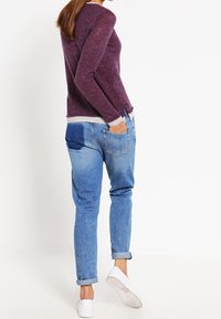 Fioletowy sweter o fakturowanej strukturze z jasnoszarym wykończeniem u dołu, zestawiony z jasnoniebieskimi dżinsami posiadającymi tylną kieszeń, podwiniętymi mankietami i białymi tenisówkami.