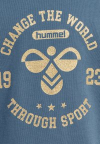 Blått tyg med en texturerad yta som har guldglittrande text och grafiska element. Texten lyder "FÖRÄNDRA VÄRLDEN GENOM SPORT" med stjärnor.