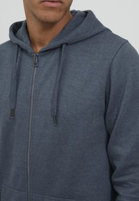 Haut zippé gris fabriqué en tissu doux, doté d'une capuche avec cordon de serrage, de poches avant et d'un zip argenté avec des tirettes foncées pour un accès facile.