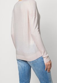 Personne portant un pull en maille à manches longues rose clair et un jean en denim bleu, vue de dos sur un fond uni.