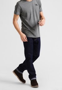 Šedé tričko s krátkým rukávem a logem, tmavě modré džíny s ohrnutými kotníky a hnědé boty s texturovaným povrchem.