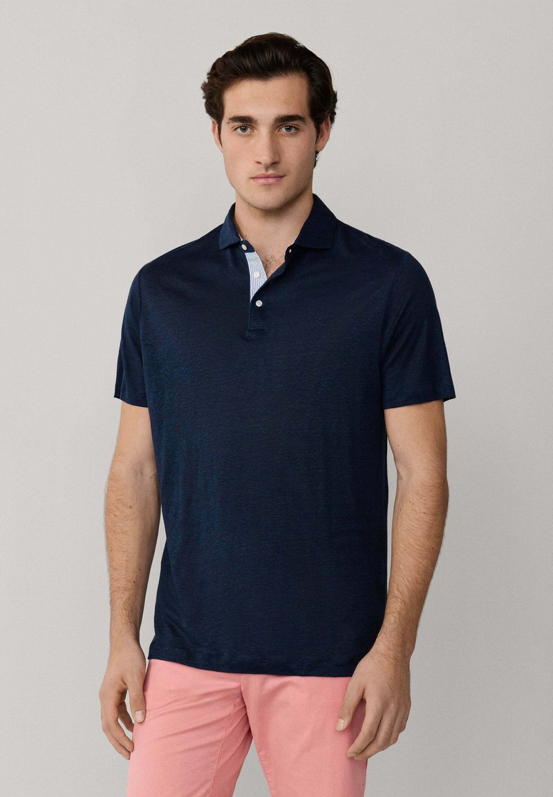 Mann, der ein marineblaues Poloshirt mit kurzen Ärmeln und korallrosafarbene Hosen trägt, steht vor einem einfarbigen hellgrauen Hintergrund.