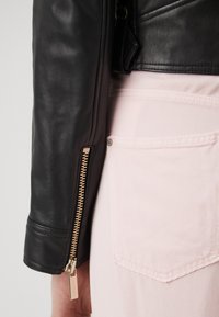 Giacca di pelle nera con dettaglio della zip dorata sulla manica, abbinata a un tessuto rosa chiaro che rivela una tasca. Texture liscia, design aderente.