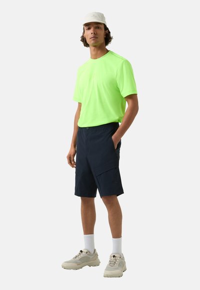Neon grøn t-shirt, marineblå cargo shorts med lommer, hvide ankelsokker, beige sneakers og en lys kasket med branding.