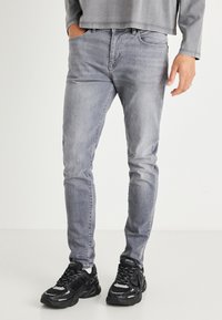 Szürke farmernadrág, slim fit fazonban, öt zsebbel és finom kopásokkal. Fekete sportcipővel párosítva.