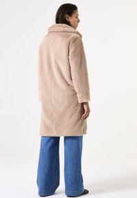 Abrigo de piel sintética beige con cuello, mangas largas y diseño hasta la rodilla, combinado con pantalones de mezclilla azul de pierna ancha. Tejido sencillo y texturizado.