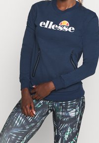 Vrouw draagt een marineblauwe ellesse-trui met ritssluitingen en patroonlegging met groene en witte bladdesigns.
