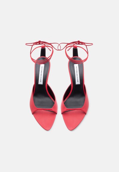 Κόκκινα σανδάλια με ψηλό τακούνι, μυτερά δάχτυλα, δεσίματα στον αστράγαλο και μαύρες σόλες με την ετικέτα Victoria Beckham, φαίνονται από πάνω σε λευκό φόντο.