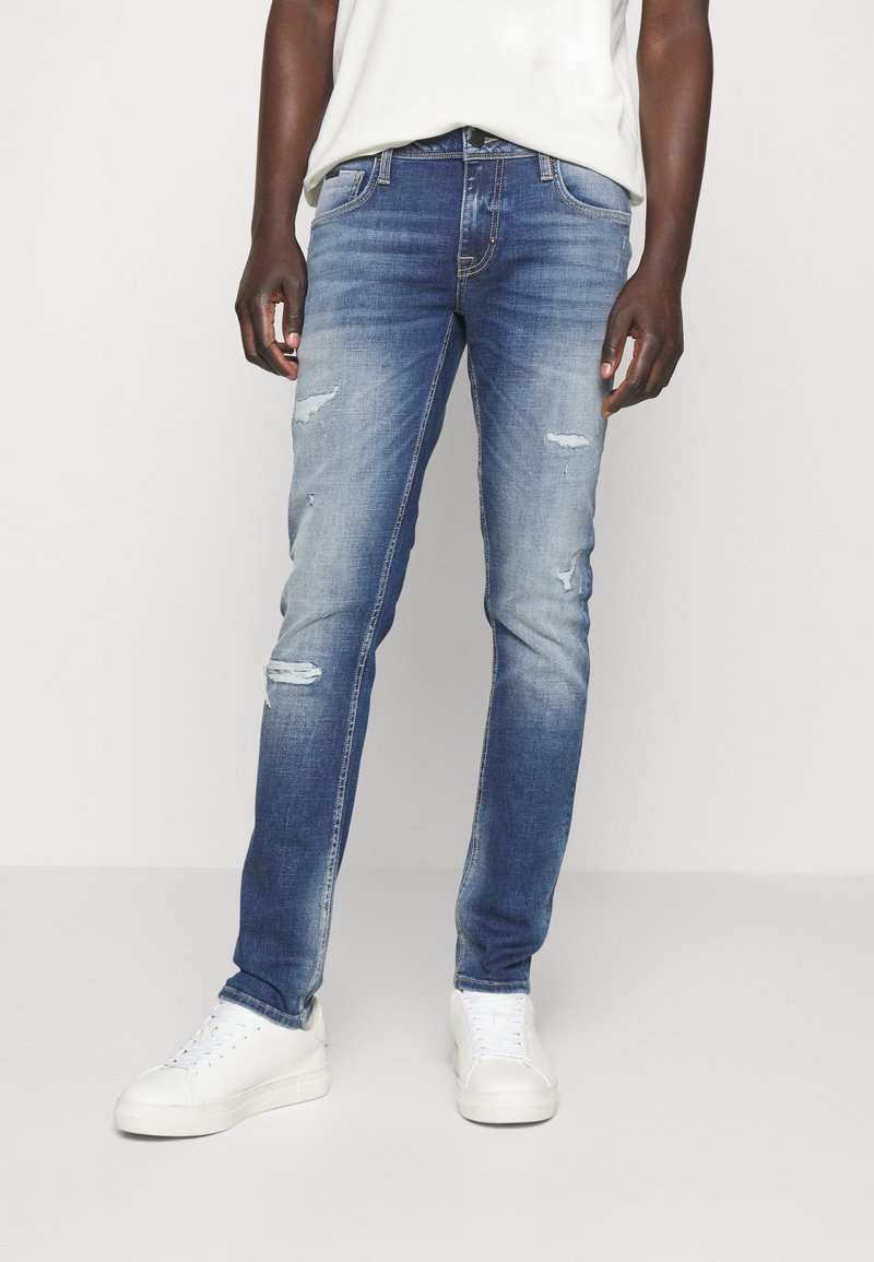 Jeans de denim azul ajustados con un ligero desgaste y detalles deshilachados. Presenta un diseño tradicional de cinco bolsillos y una cremallera con cierre de botón.