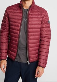 Bordeauxrote gesteppte Daunenjacke mit durchgehendem Reißverschluss, gerippten Bündchen und horizontalen Nähten. Wird über einem grauen T-Shirt getragen, das ein rundes Logo auf der Brust zeigt.