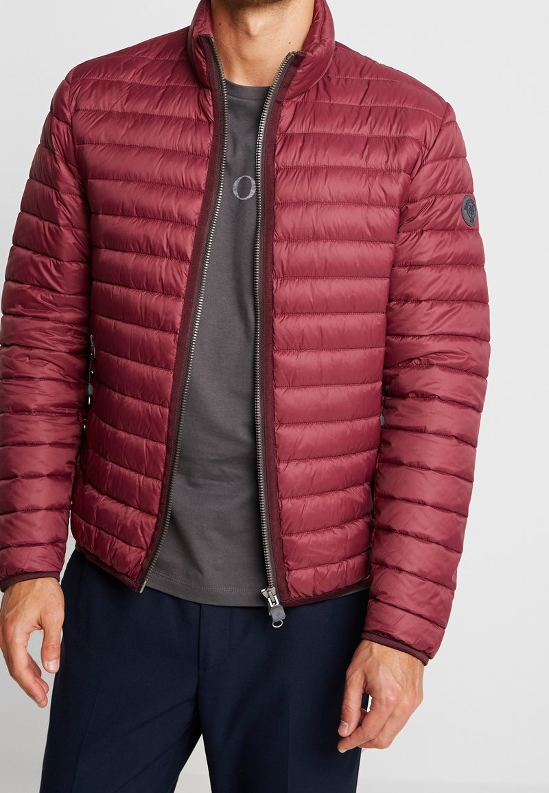 Bordeauxrote gesteppte Daunenjacke mit durchgehendem Reißverschluss, gerippten Bündchen und horizontalen Nähten. Wird über einem grauen T-Shirt getragen, das ein rundes Logo auf der Brust zeigt.