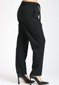 Donna che indossa pantaloni neri a vita alta a gamba stretta con elastico in vita e scarpe col tacco alto a punta nera su sfondo bianco.