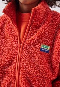 Dziecko w jaskrawo pomarańczowej, teksturowanej polarowej kurtce z zamkiem błyskawicznym i naszywką "La Maison Amy Super Vintage" na piersi.