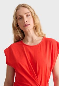 Haut rouge à manches courtes en tissu doux, présentant un col rond et un design torsadé à la taille pour une touche de détail supplémentaire.