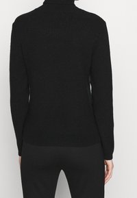 Personne portant un pull col roulé noir côtelé ajusté et un pantalon noir, vue de dos sur un fond blanc uni.