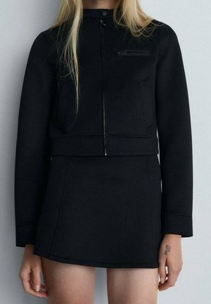 Kobieta ubrana w czarną kurtkę na zamek i dopasowaną czarną mini spódnicę, stojąca na tle jasnego, jednolitego tła, z widocznym małym tatuażem na dłoni.