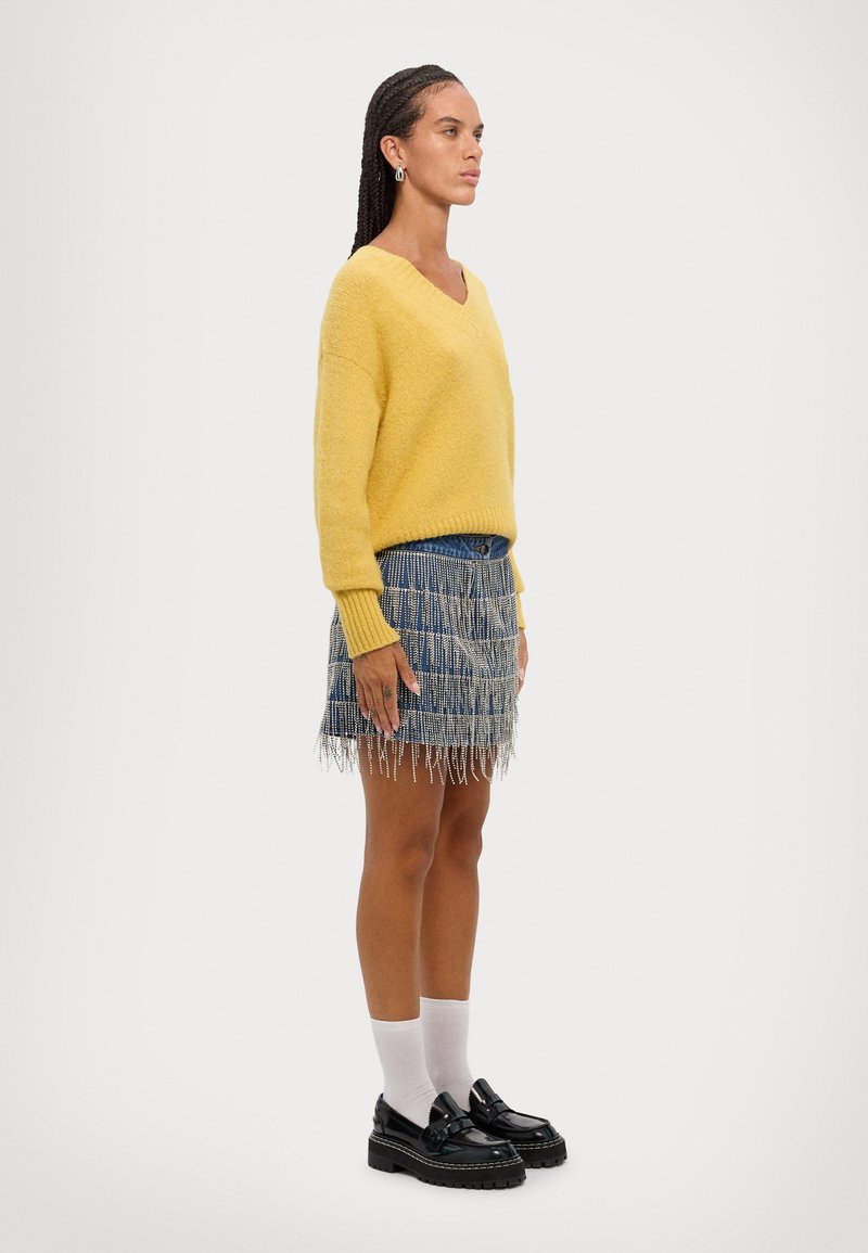 Gelber gestrickter Pullover mit V-Ausschnitt, kombiniert mit einem Jeans-Minirock, der mit vertikalen Fransen verziert ist. Schwarze Slipper und weiße Socken vervollständigen den Look.