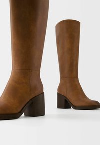 Bruna läderstövlar i knähöjd med en kraftig klack, slät yta och minimalistisk design, med rund tå och enkla sömdetaljer.