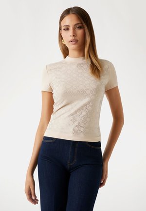 Top beige a maniche corte con un motivo a diamante strutturato, collo alto e vestibilità aderente. Abbinato a pantaloni in denim blu scuro.