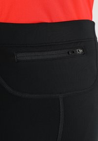 Černé sportovní kalhoty s kapsou na zip na zadní straně, hladkým povrchem a prošívanými detaily podél švů, v kombinaci s červeným topem.