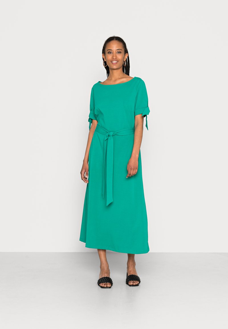 Vestido midi em teal com mangas curtas dobradas, um cinto atado à cintura e um corte relaxado. Combinado com sandálias tipo slide pretas texturizadas.