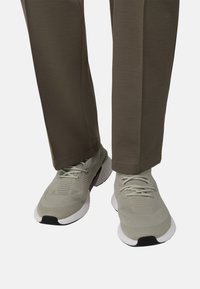 Chaussures de sport gris clair avec une tige en mesh texturé, une semelle en caoutchouc blanche, et un design flexible et rembourré, associées à un pantalon vert olive.