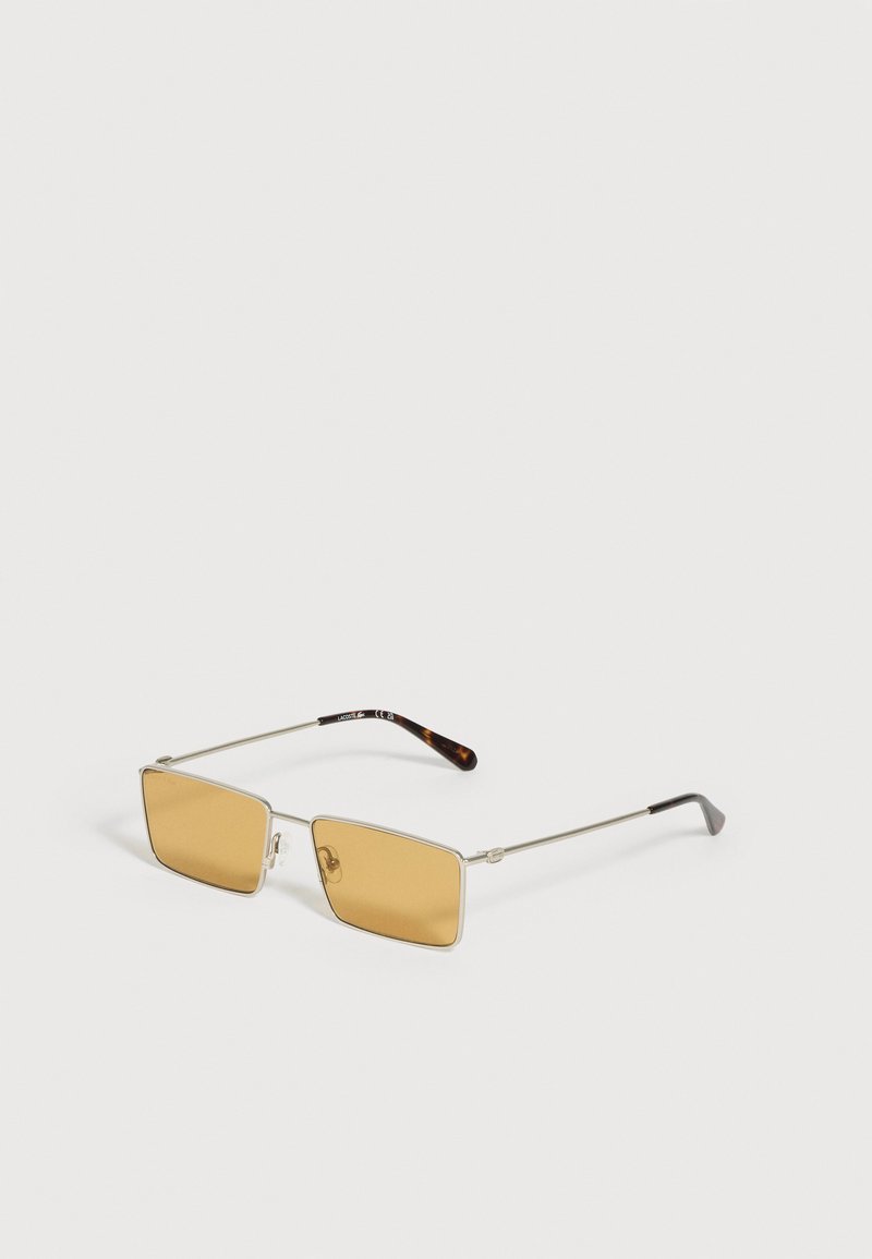 Lunettes de soleil rectangulaires avec fines montures métalliques argentées et verres teintés ambre, reposant sur une surface blanche.
