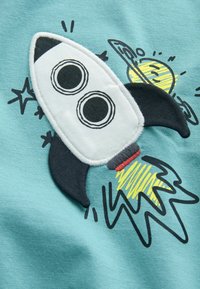 Turkusowy materiał zdobi haftowana rakieta o białej sylwetce, czarnych płetwach i dwóch dużych okrągłych oknach, otoczona gwiazdami i planetą.