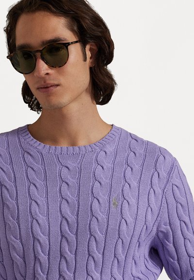 Μωβ πουλόβερ με πλέξη καλωδίων και στρογγυλή λαιμόκοψη, διακοσμημένο με ένα μικρό κεντημένο λογότυπο. Συνοδεύεται από σκούρα γυαλιά ηλίου από ταρταρούγα.