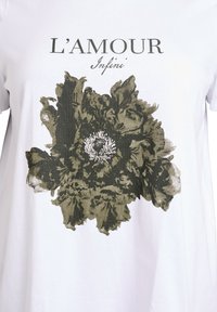 Bílá tričko s velkým tmavě zeleným květinovým potiskem uprostřed a textem "L'AMOUR Infini" nad designem.