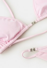 Bikini rose clair avec bords froncés et attaches papillon argentées sur les bretelles, posé à plat sur un fond blanc.