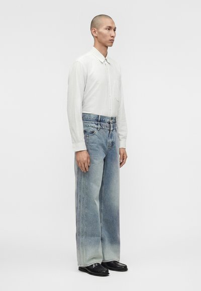 Άνδρας που φορά λευκό πουκάμισο με μακριά μανίκια, ξεθωριασμένο φαρδύ τζιν με ορατές ραφές και μαύρα μοκασίνια, σε μονόχρωμο ανοιχτό φόντο.
