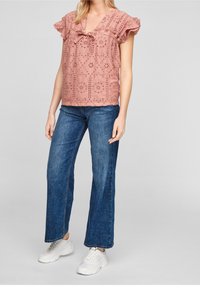 Blusa rosa con design in pizzo floreale e maniche a volant, abbinata a jeans blu a gamba dritta e sneaker bianche.