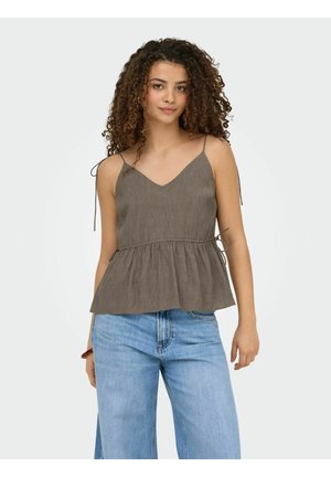 Femme aux cheveux bouclés portant un haut marron sans manches avec des liens aux épaules et à la taille, associé à un jean bleu clair, debout sur un fond blanc.