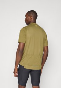 Olivegrünes Sporthemd mit kurzen Ärmeln und strukturiertem Finish, mit einer Rückenentasche und reflektierenden Details am Saum. Schwarze enganliegende Shorts.