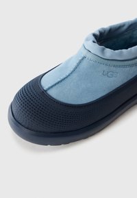 Světle modré semišové UGG slip-on boty s polstrovaným límcem a texturovanou námořnickou gumovou špičkou a podrážkou na bílém pozadí.