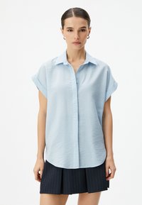 Camisa de botones de manga corta en azul claro con un acabado texturizado, cuello clásico y dobladillo curvado. Combinada con una falda oscura plegada.